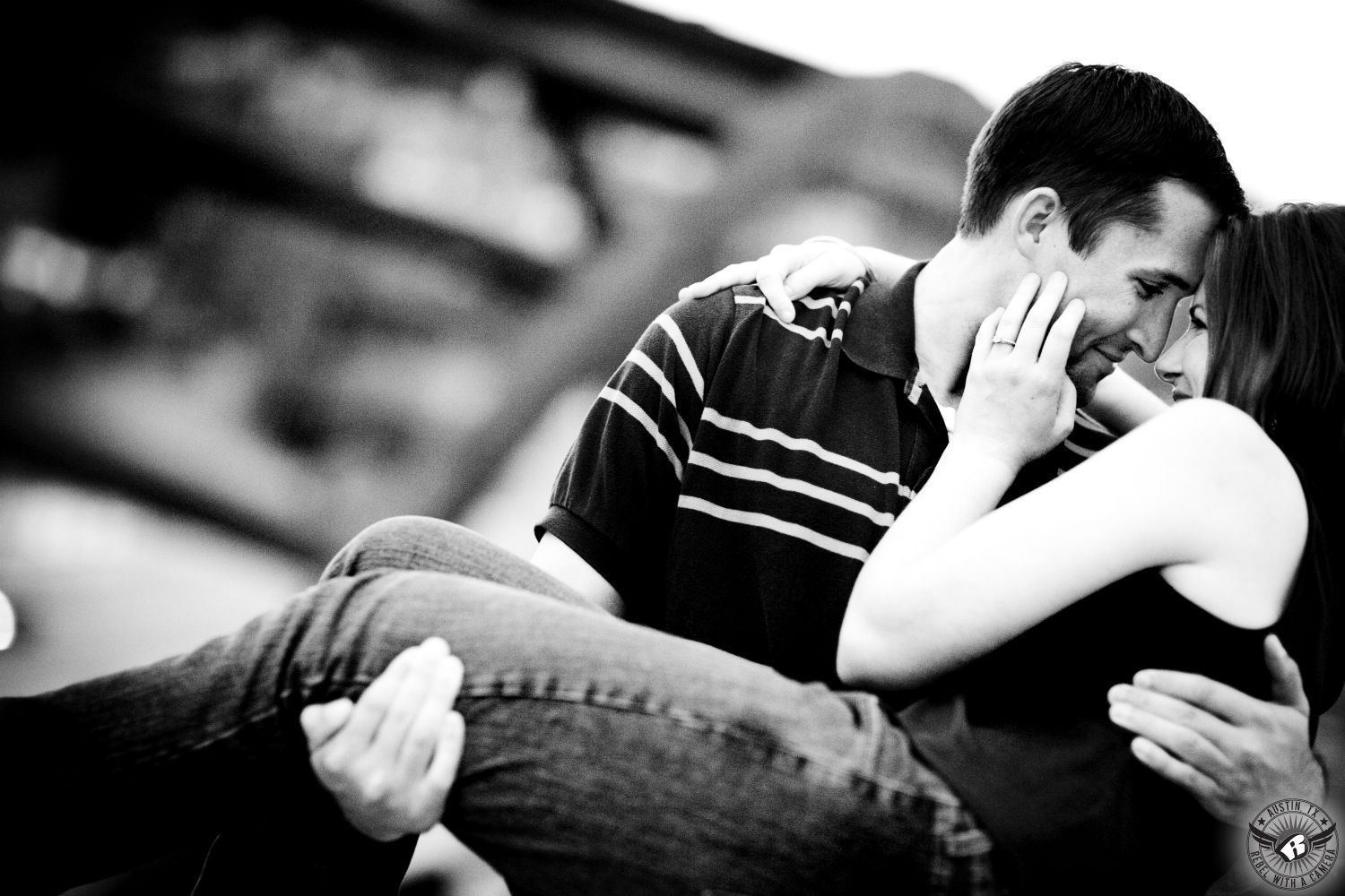 Dark haired girl in a black tank top and dark jeans touches the cheek and smiles at a dark haired guy wearing a dark shirt with white stripes who is picking her up in front of the Pennybacker Bridge in the passionate engagement photo at the 360 Bridge overlook in Austin Texas.